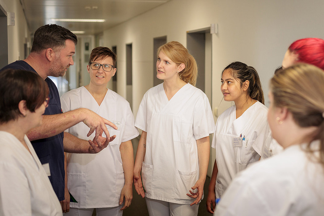 Gruppe von sechs Personen in weißen Kitteln steht im Flur und hört einem Mann zu, der mit Händen gestikuliert