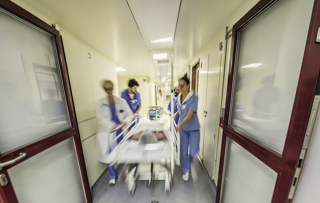Krankenhausflur mit drei medizinischem Personal in blauer Kleidung, die eine liegende Person auf einer Trage schieben