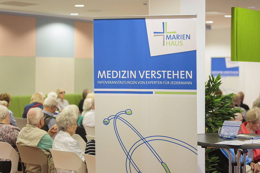 Rollup mit dem Schriftzug 'MEDIZIN VERSTEHEN INFOVERANSTALTUNGEN VON EXPERTEN FÜR JEDERMANN' und dem Logo 'MARIEN HAUS' vor einem Publikum in einem Veranstaltungsraum