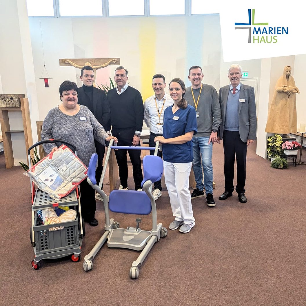 Gruppe von sieben Personen in einem Raum mit Marienhaus-Logo, medizinischem Hebehilfe-Gerät und Statue im Hintergrund