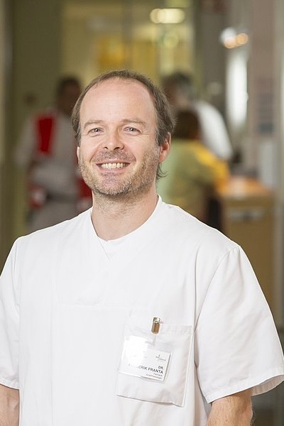 Mann in weißer medizinischer Arbeitskleidung mit Namensschild in einem Krankenhausflur